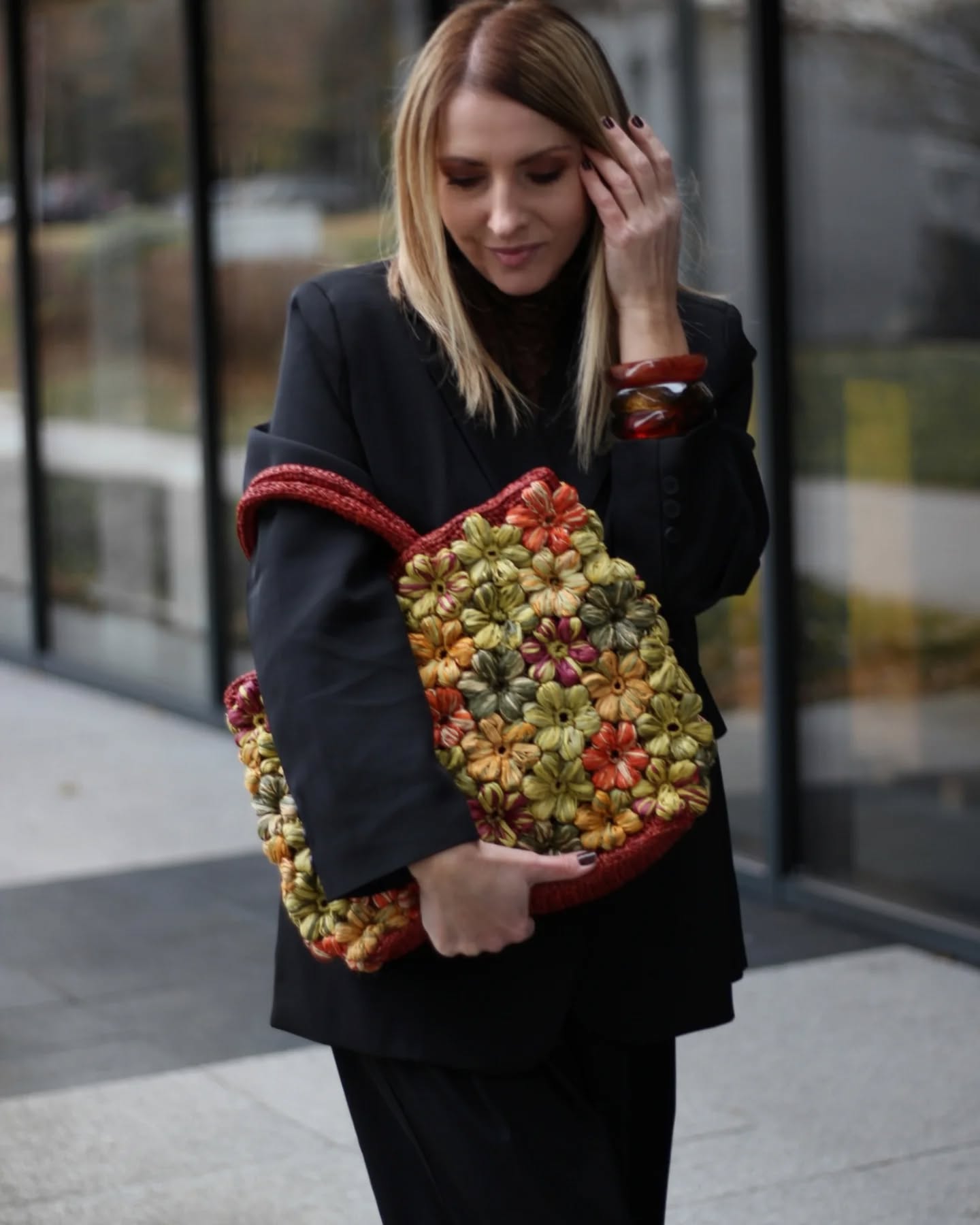 a women in black outfit holding a floral crochet raffia bag in hand, the floral bag is in a red base color with green, yellow, red, orange tone puff flowers