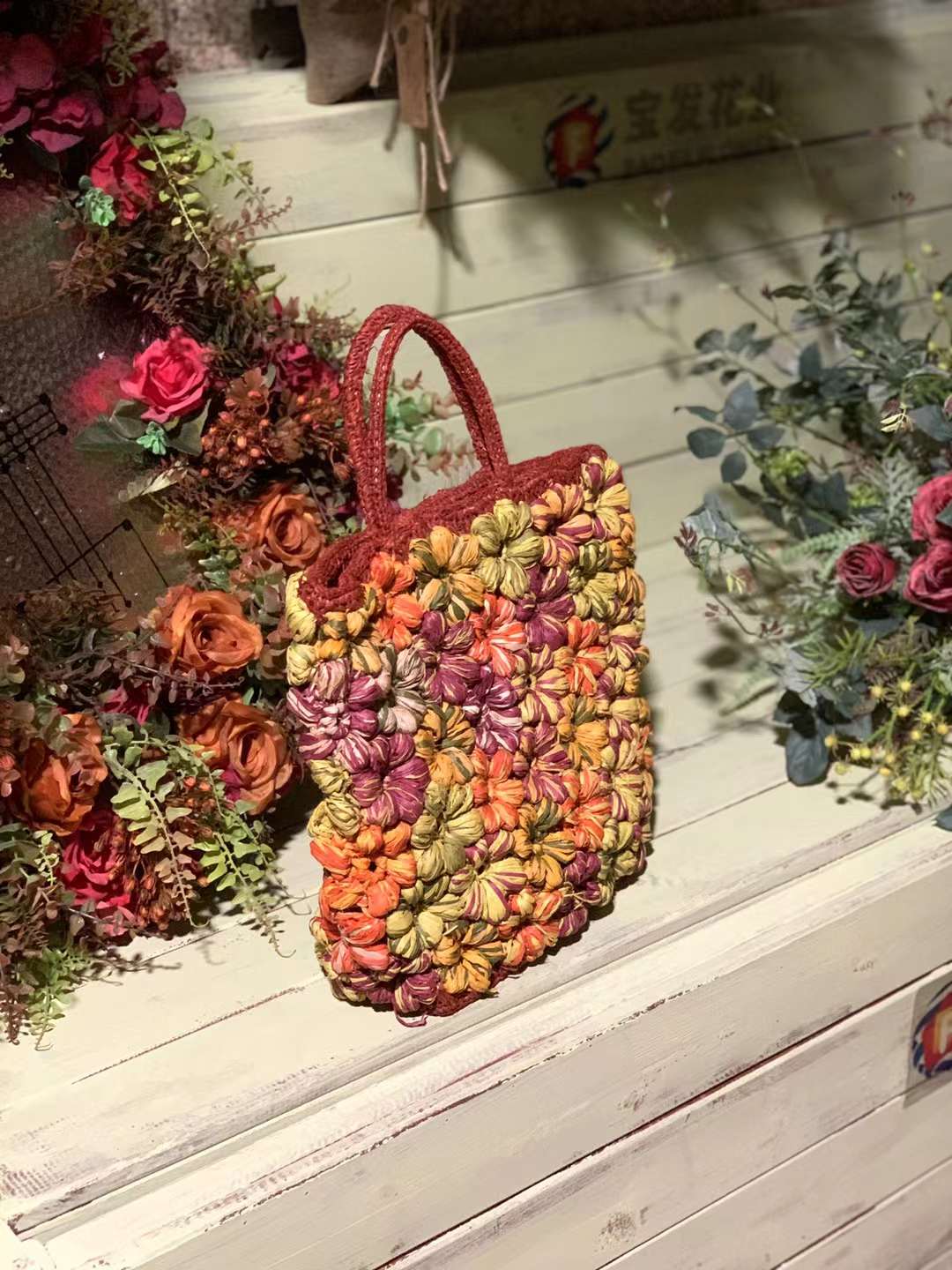 A handmade red straw bag with flowers design. The bag is in a red color base adorned with orange, yellow, and pink crochet flowers. The bag has two top handles and a flat bottom, and it is made in natural straws.