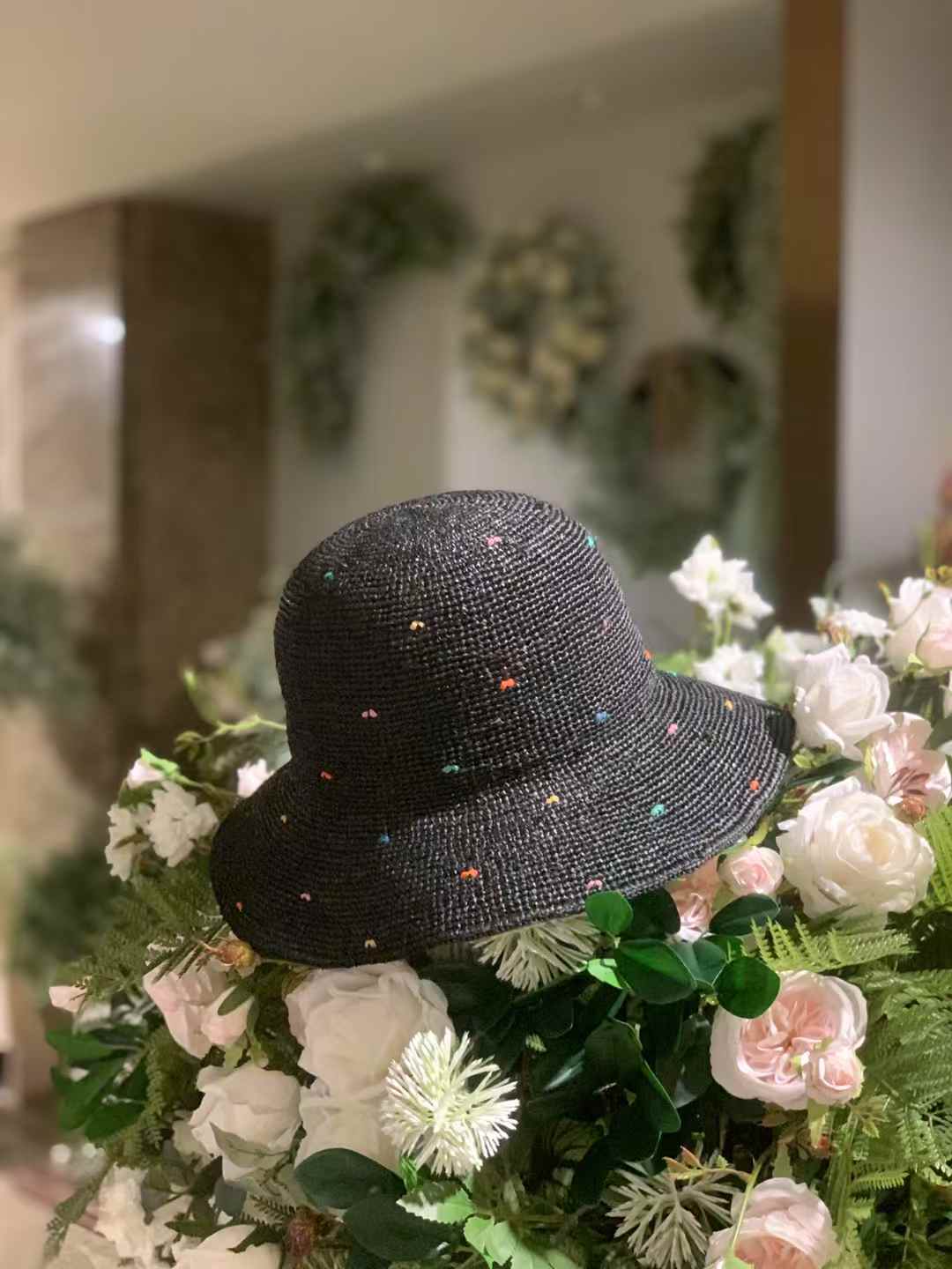 Black color raffia straw hat with wide brim and colorful speckles, hand-crocheted sun hat displayed on a bunch of artificial flowers