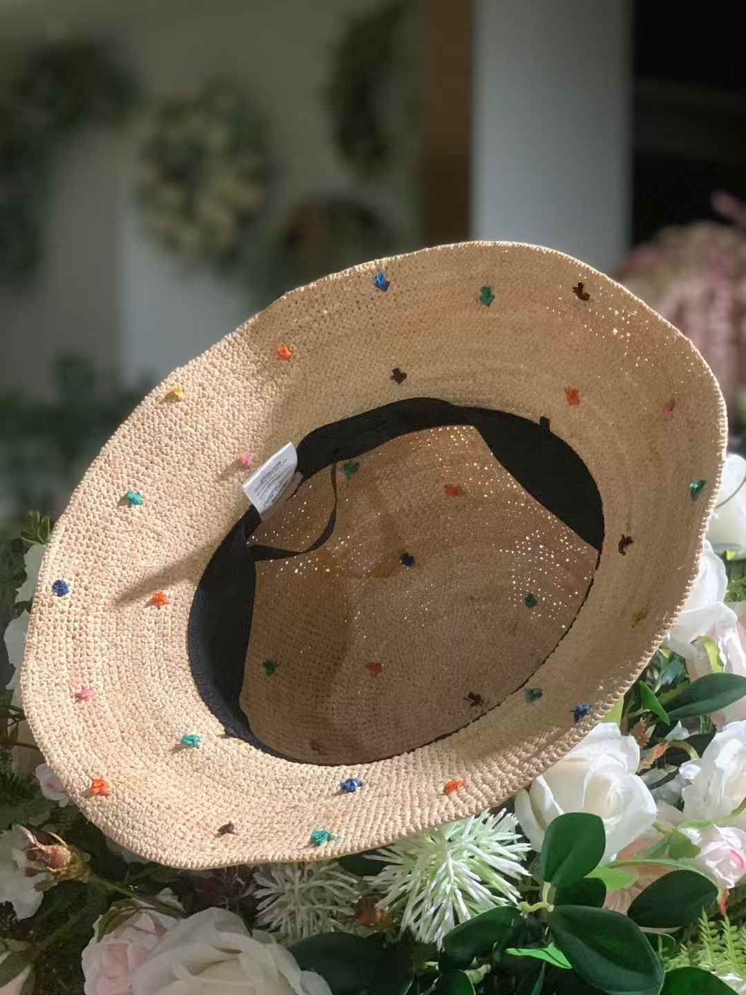 the inner vision of a natural beige raffia straw hat with wide brim and colorful speckles, with a black adjustable band, laying on a bunch of artificial flowers