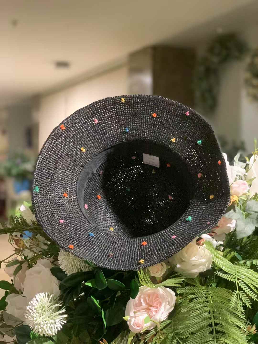 a black color raffia straw hat with wide brim and colorful speckles, up side down displayed on a bunch of artificial flowers, stylish sun hat for sea side