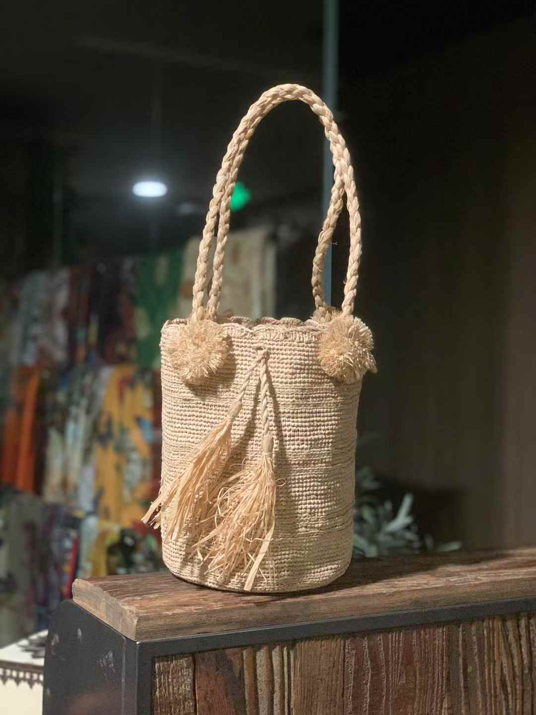 drawstring raffia bucket bag in earth tone beige color with pom pom on the top of edge, 2 shoulder straps