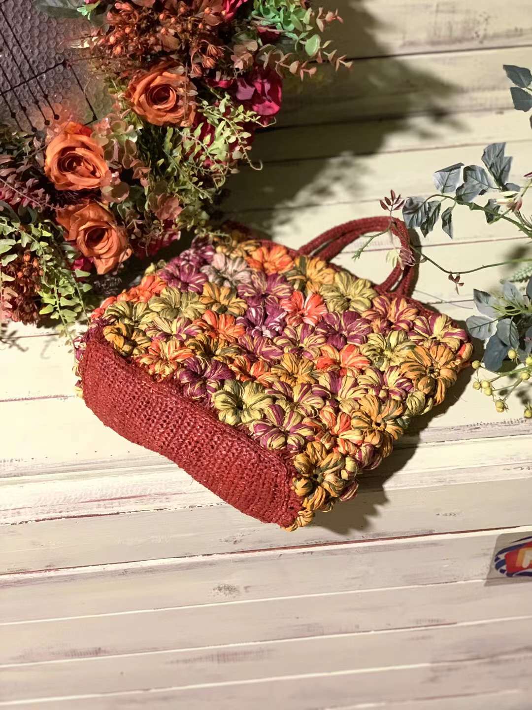 A crocheted tote bag with a floral design. The bag is made of colorful yarn in shades of pink, orange, yellow, and green. The bag has a brown handle and is sitting on a white wooden surface.