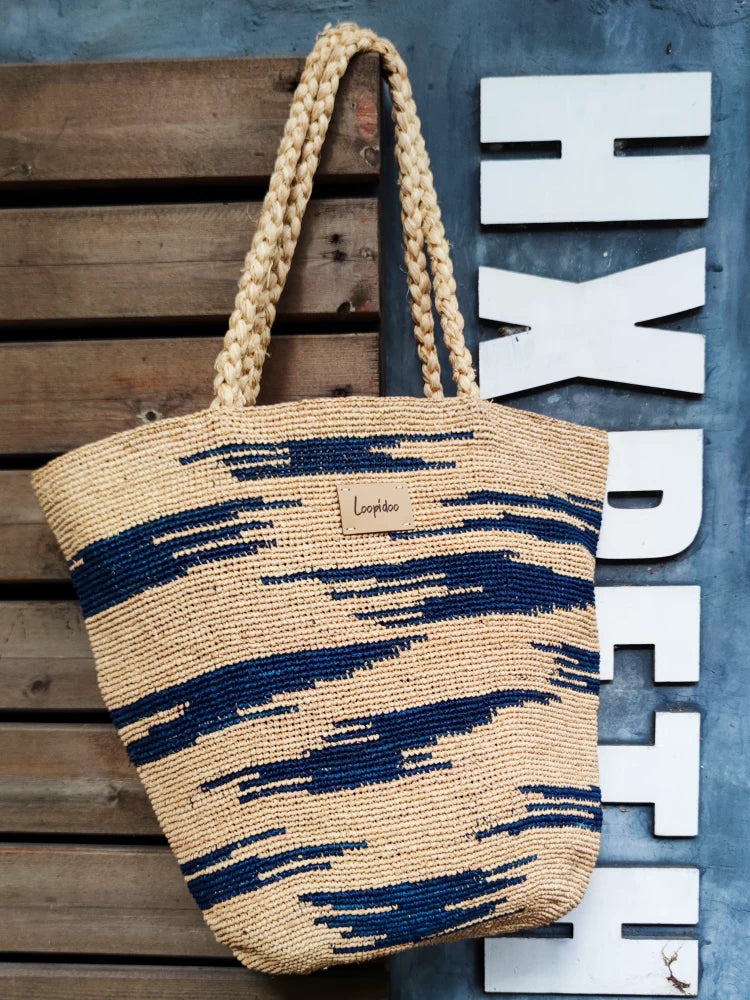 a raffia bag in beige color base, with navy color camouflage pattern around the body. The shoulder bag is hand crochet in natural raffia straw, plat tote shape, with 2 hand woven shoulder straps in same materials hanging on a wooden wall