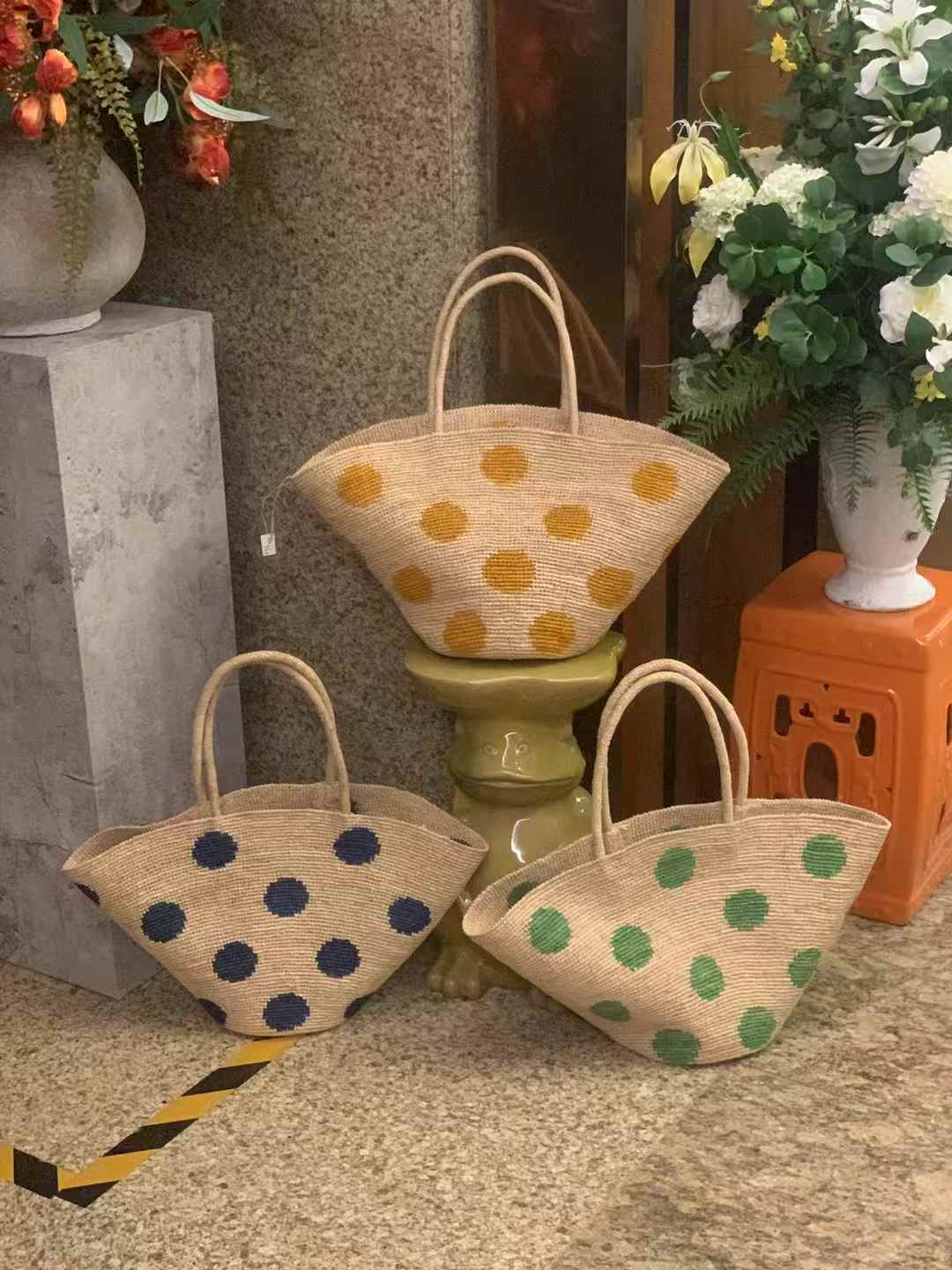 3 different colors raffia tote bag with natural base and large polka dots, each is in blue dots, green dots and yellow dots. The tote bag is in wide trapezoid shape with round bottom and raffia handles.