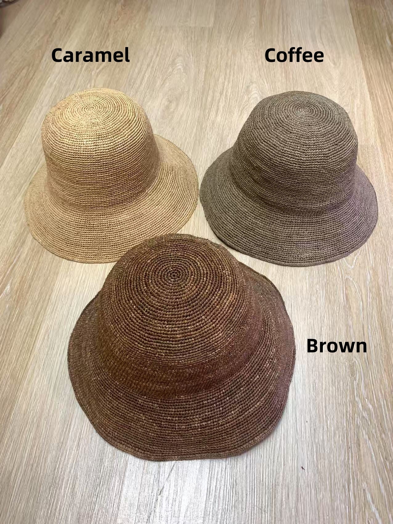3 raffia bucket hat displaying on the wooden floor, they are in caramel color, coffee color and brown color. these raffia hats are hand crochet in plain minimalist bucket hat style