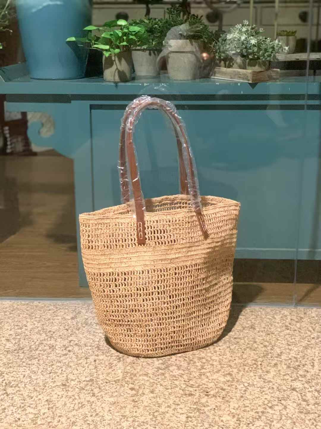 Handmade raffia tote bag with leather shoulder straps, featuring an open-loop crochet pattern in natural beige, standing upright on a stone floor.