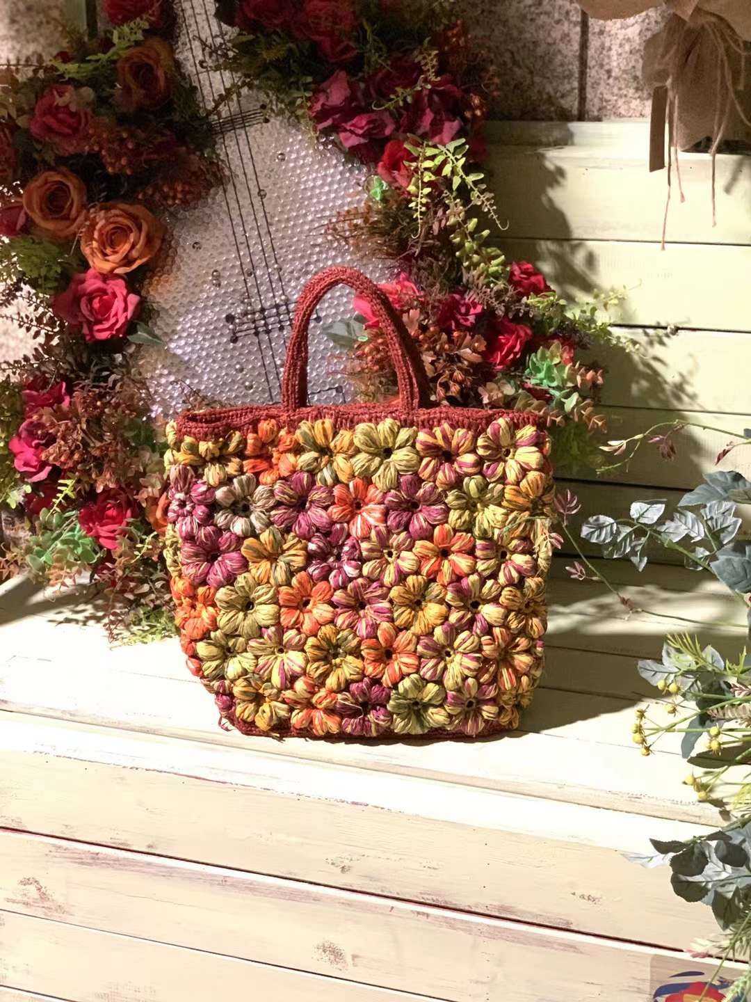 A handmade red tote bag with a floral pattern in multiple colors includes orange, yellow, green, and purple. The bag has a red brown base and two handles, with flat bottom that standing up right on a white wooden surface.