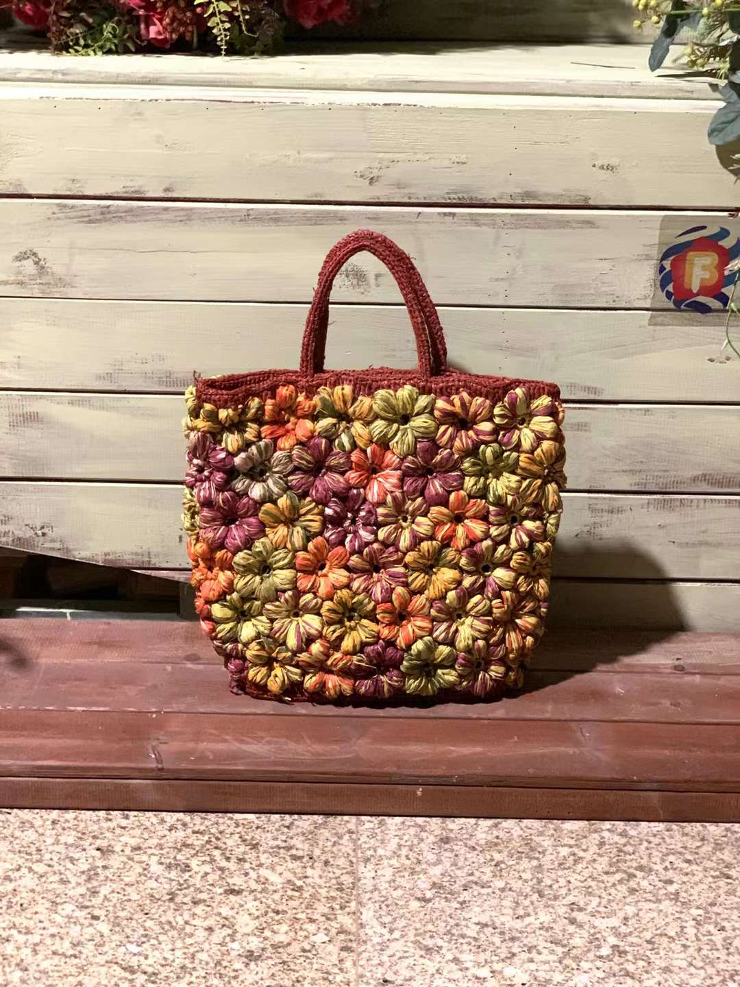 A raffia tote bag with a floral design. The bag is in a red color base adorned with orange, yellow, and pink crochet flowers. The bag has two top handles and a flat bottom.