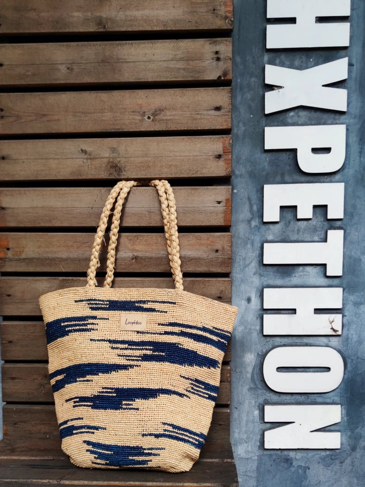 a shoulder bag in beige color base, with navy color camouflage pattern around the body. The shoulder bag is hand crochet in natural raffia straw, plat tote shape, with 2 hand woven shoulder straps in same materials hanging on a wooden wall