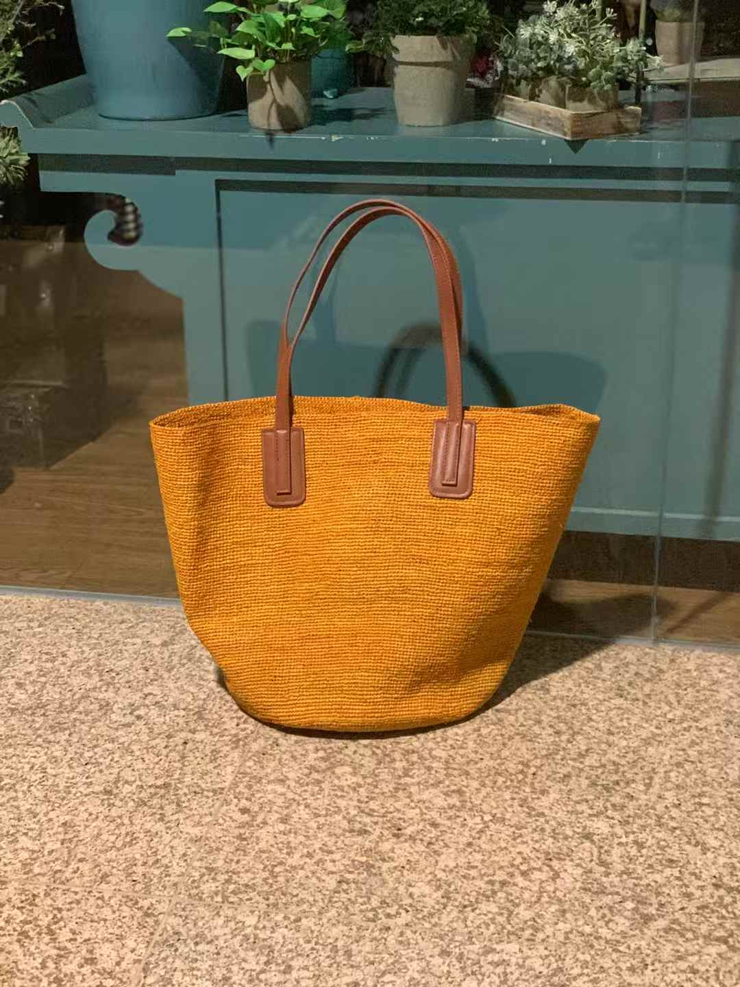 an orange color large straw bag with brown color leather shoulder strap, solid color in plain pattern, wide opeing, the tote bag is standing upright on a marble floor.