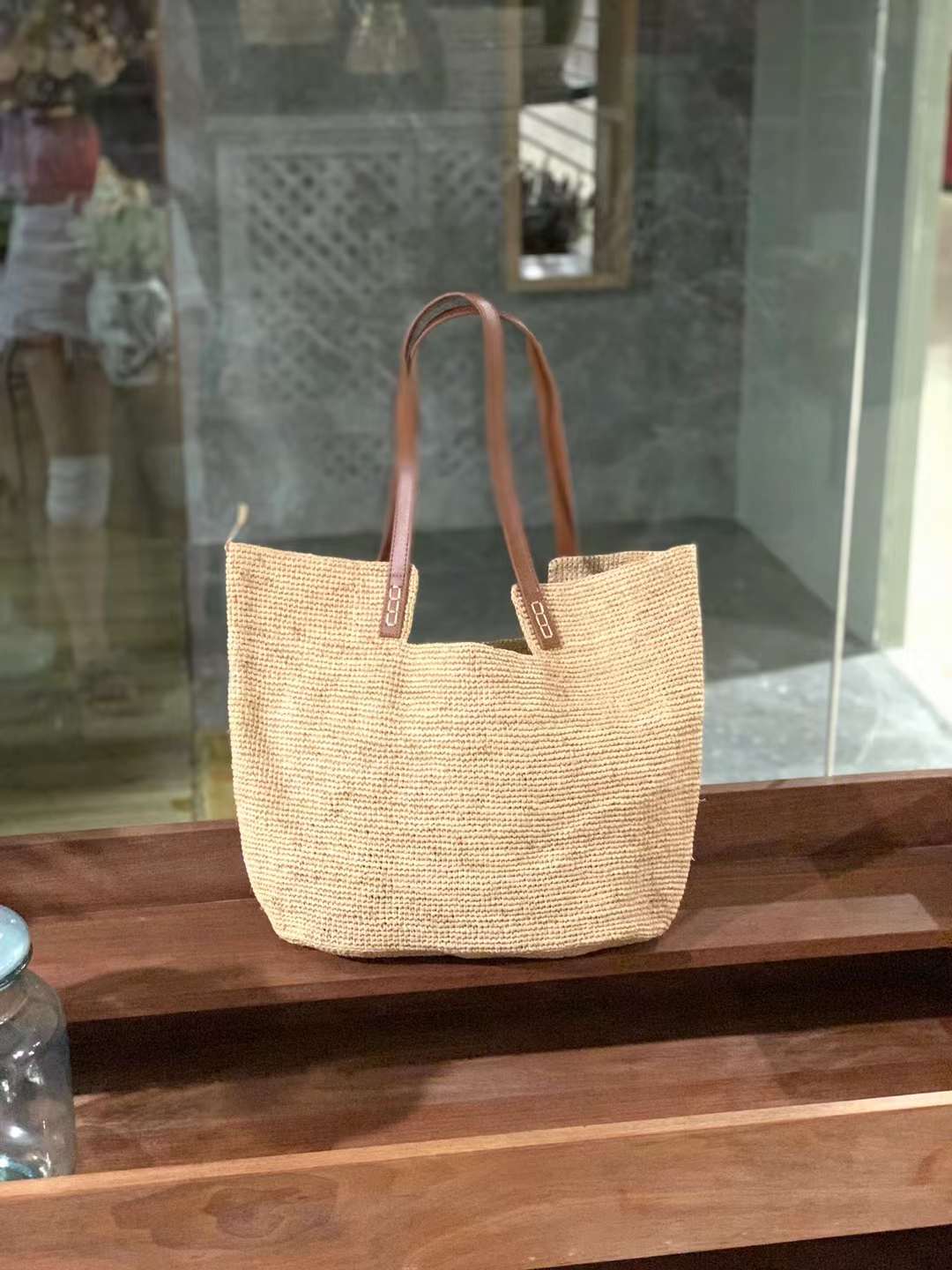 A minimalist hand crochet straw tote in square flat shape, natural beige with brown leather shoulder strap, the straw tote is display on a wooden table