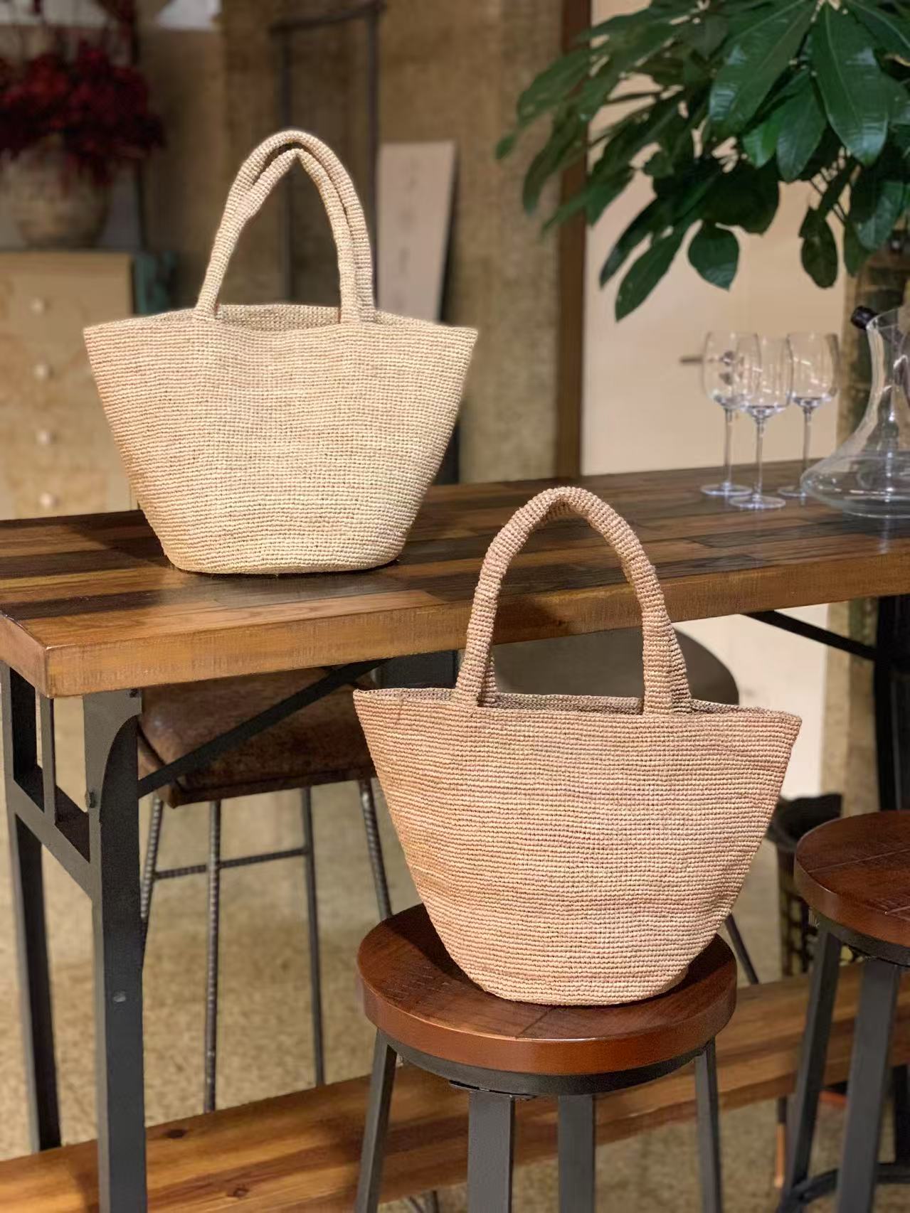 2 natural color raffia bag with top handles, one in beige color and the other one in brown color, the raffia bag is in a tote shape with round bottom, plain body and wide opening. they are displaying on wooden table and a wooden chair.