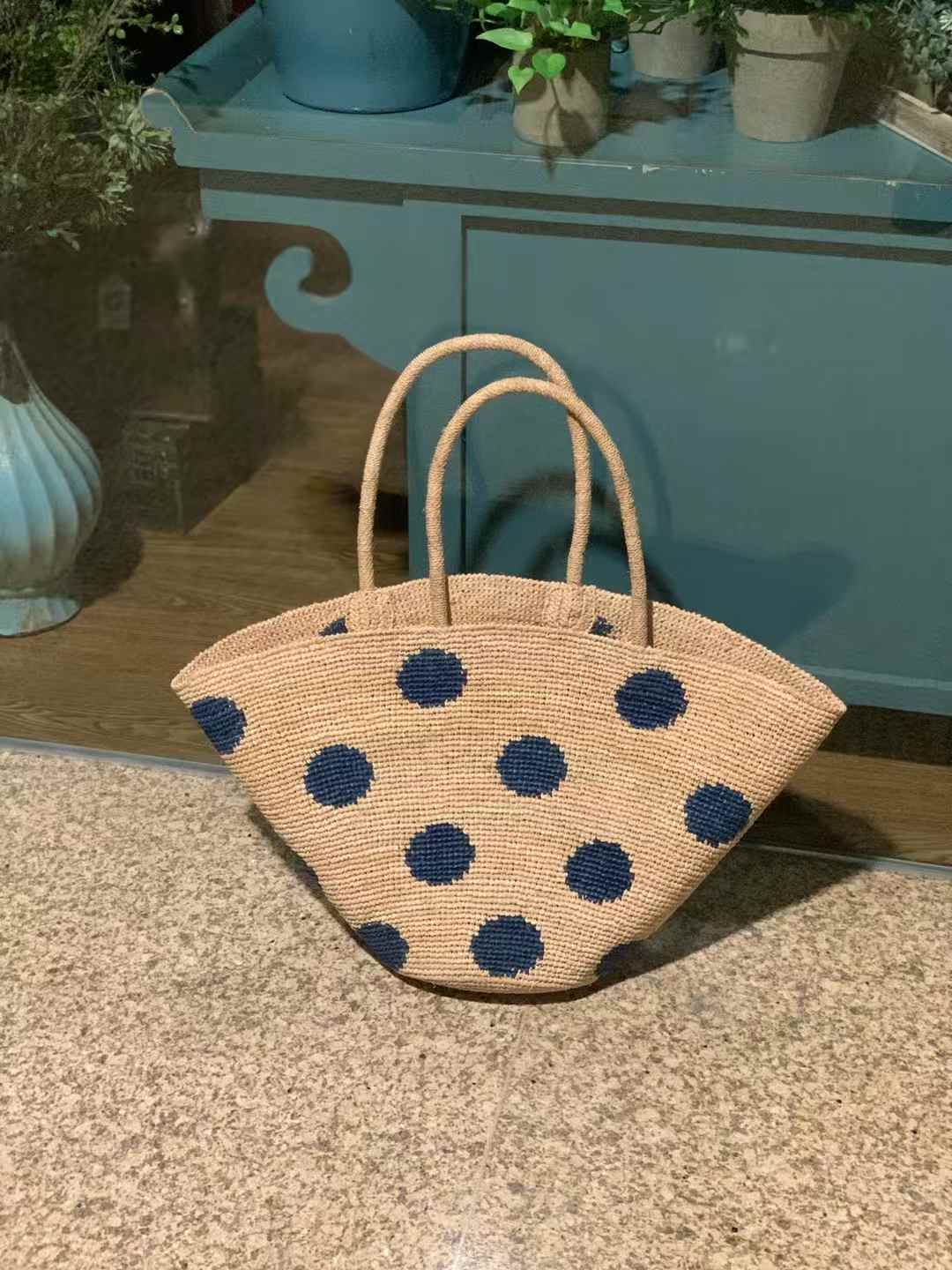 Hand-crocheted raffia bag with natural base and large blue polka dots, wide trapezoid shape with round bottom and hand woven raffia handles, this raffia tote bag is standing upright on the marble floor