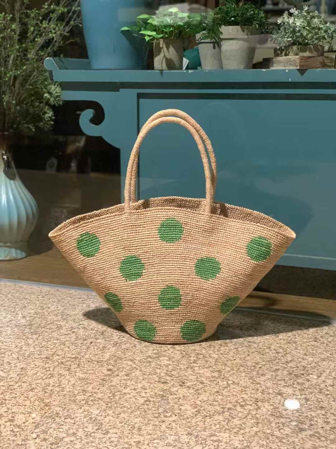 Hand-crocheted raffia bag with natural base and large green polka dots, wide trapezoid shape with round bottom and raffia handles.
