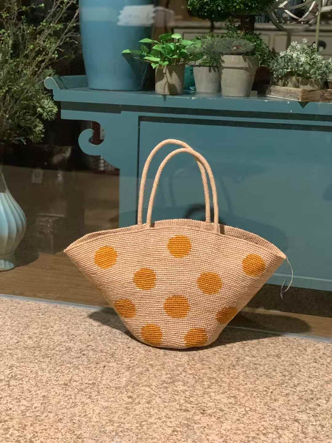 Hand-crocheted raffia tote bag with natural base and large yellow polka dots, wide trapezoid shape with round bottom and raffia handles.