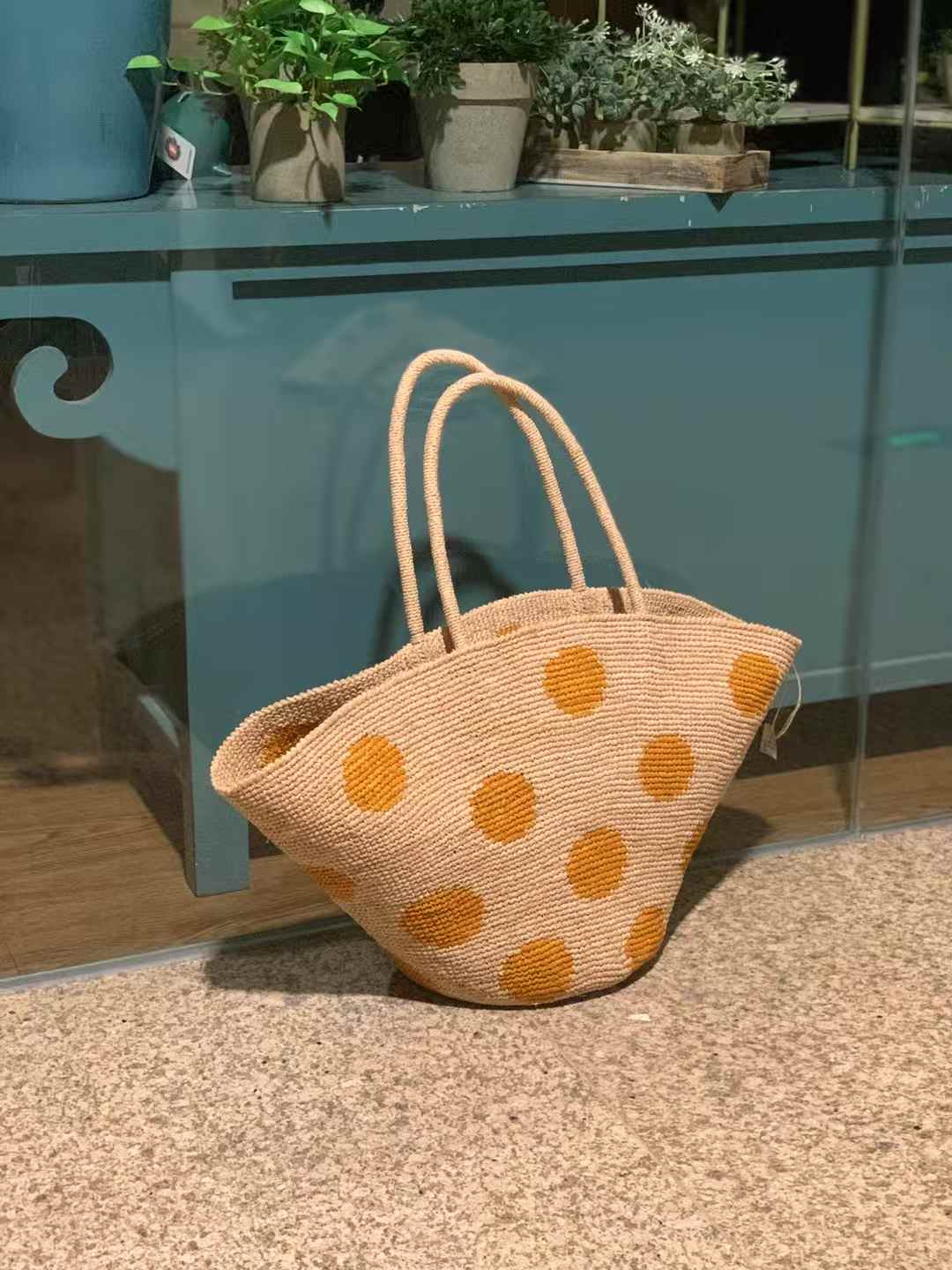 Hand-crocheted raffia tote bag with natural base and large yellow polka dots, wide trapezoid shape with round bottom and raffia handles.
