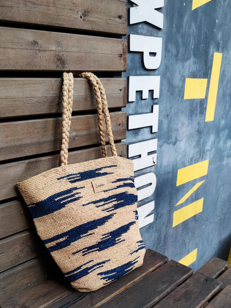 side vision of a raffia bag in beige color base, with navy color camouflage pattern around the body. The raffia bag is hand crochet in natural raffia straw, plat tote shape, with 2 hand woven shoulder straps displaying on a wooden stand