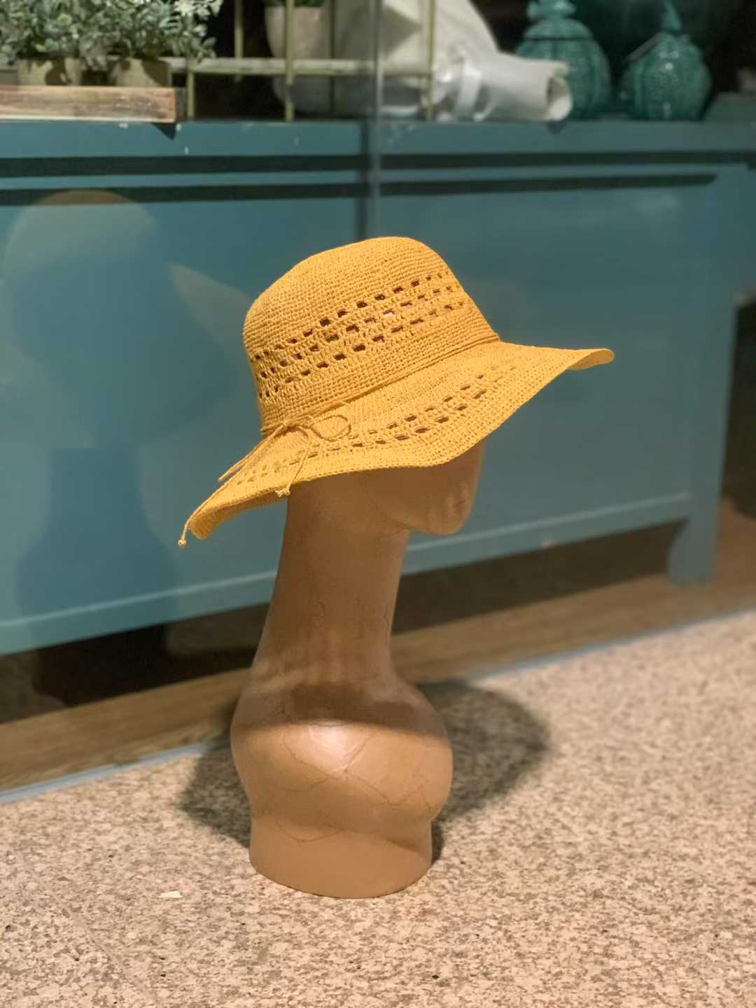 a mustard yellow color straw hat display on some artificial flowers, this summer hat has a wide brim, with hollow crochet pattern, with a straw string tie in a bow, design in floppy style for women, and it is hand handmade in raffia straw